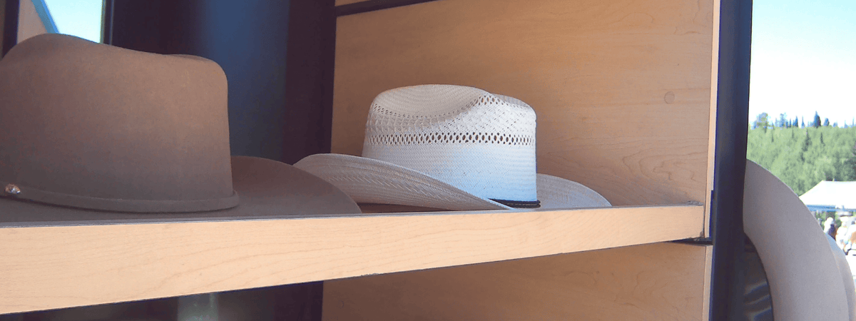 A close-up of several cowboy hats displayed on light wood shelves, including a tan wide-brim hat, a white woven-brim hat, and a cream hat partially visible on the right at a custom hat bar event.