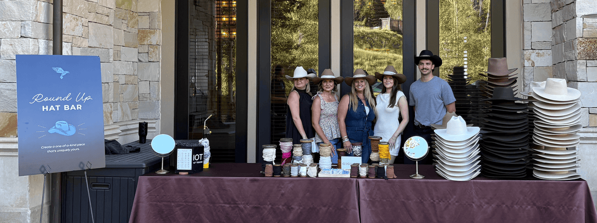 A group of five people standing behind a table at a custom hat bar event, with stacks of white and black cowboy hats on either side. A blue sign on the left reads 'Round Up Hat Bar'.