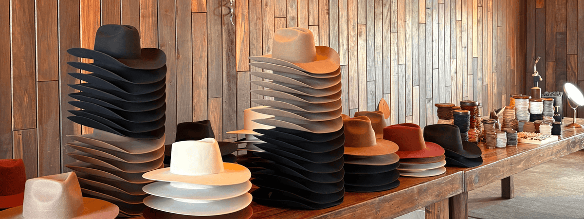 Stacks of various cowboy and western hats in black, beige, brown, and red displayed on a wooden table against a wood-paneled wall at a custom hat bar event.