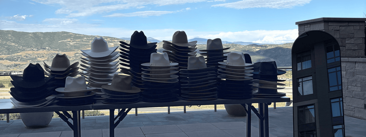 Stacks of cowboy hats in black, white, and tan displayed on folding tables outdoors with a scenic mountain landscape and a stone building in the background at a custom hat bar event.