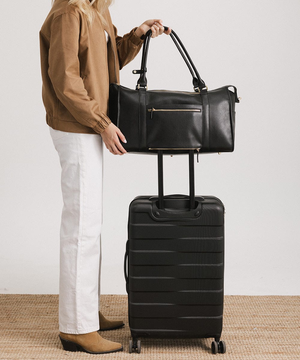 Person wearing a brown jacket, white pants and brown boots, placing a black vegan leather weekender bag on a black suitcase against a plain background #color_black