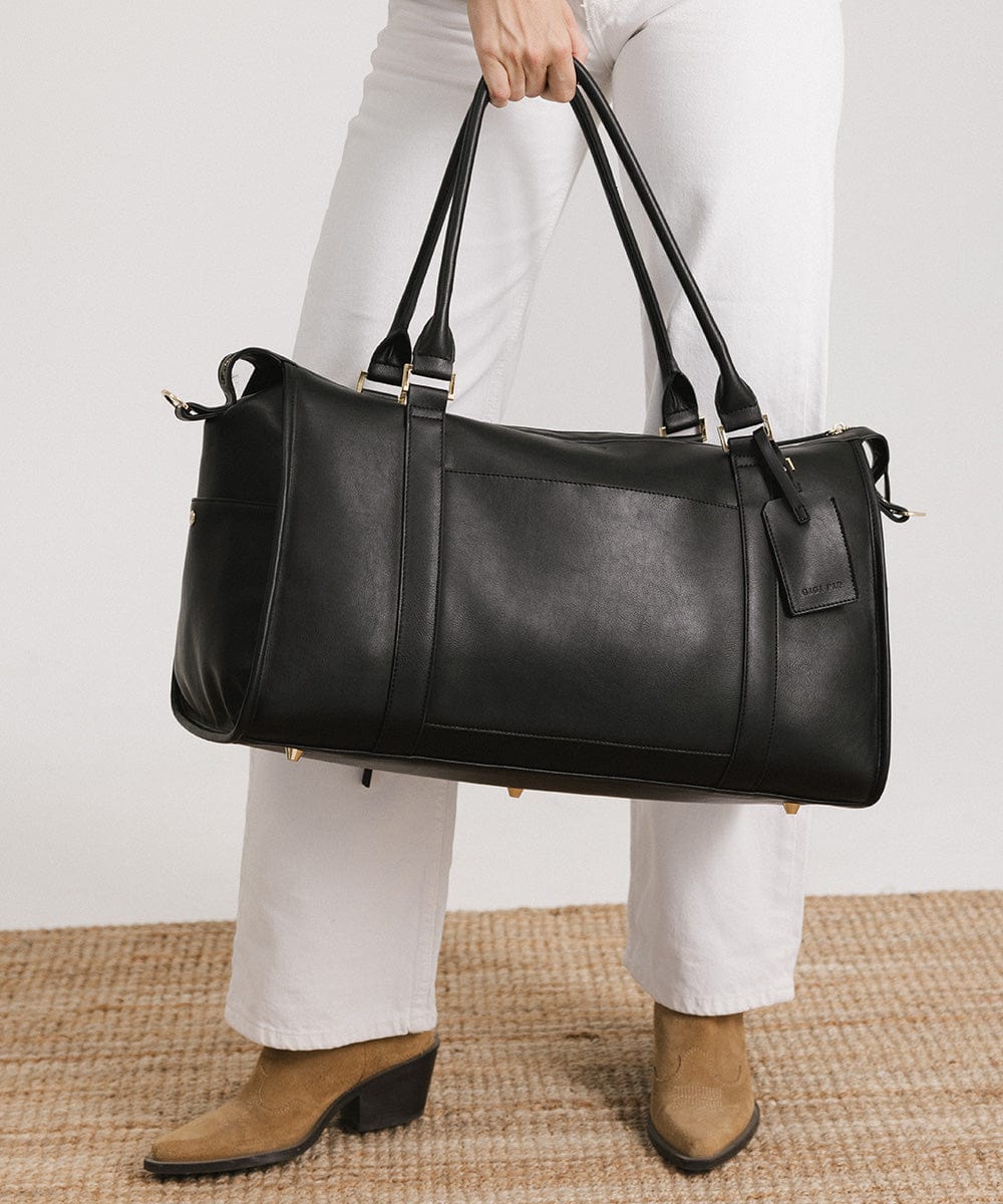 Person wearing a white pants and brown shoes carrying a black vegan leather weekender bag against a plain background #color_black
