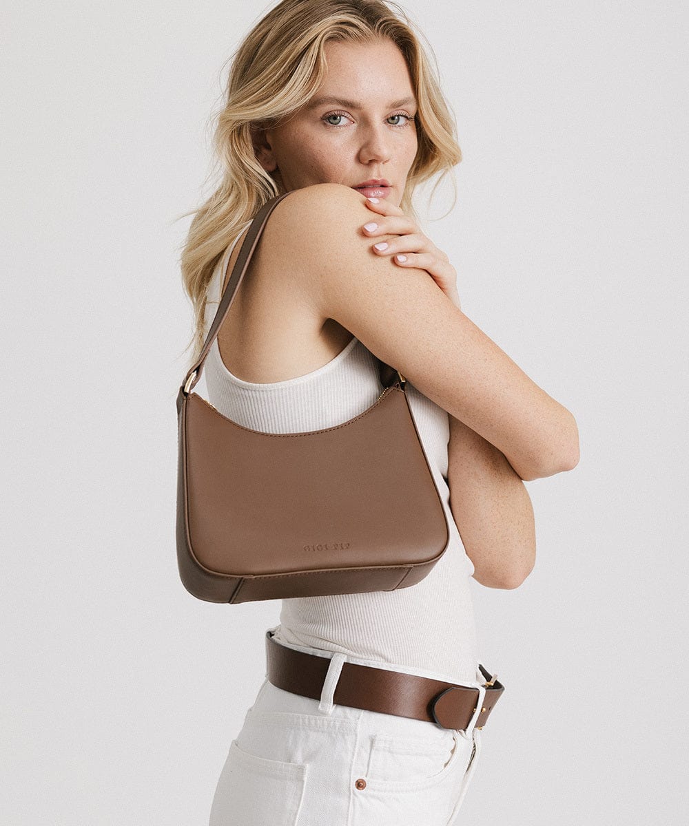 Woman wearing a white top, carrying a chocolate colored genuine leather handbag against a plain background #color_chocolate
