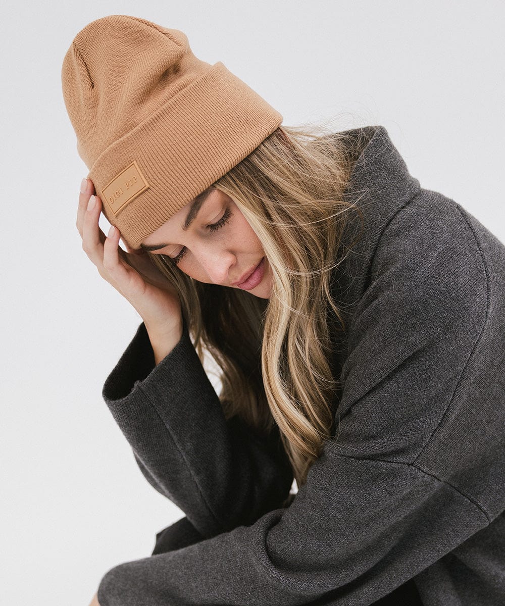 Woman wearing a latte colored knit beanie and a grey sweater, posing sideways against a plain background #color_latte
