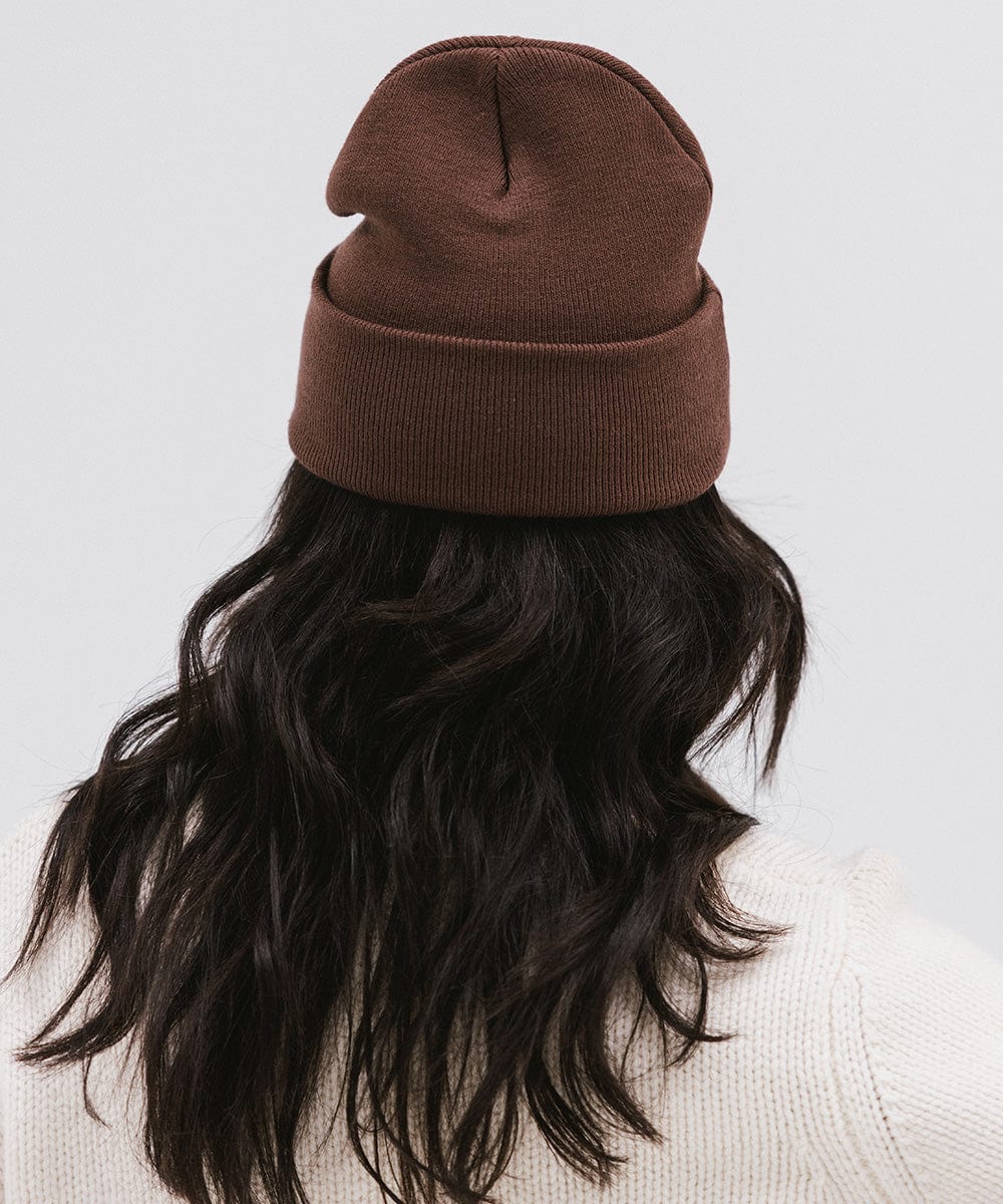 Woman wearing a chocolate colored knit beanie and a white sweater, facing behind against a plain background #color_chocolate