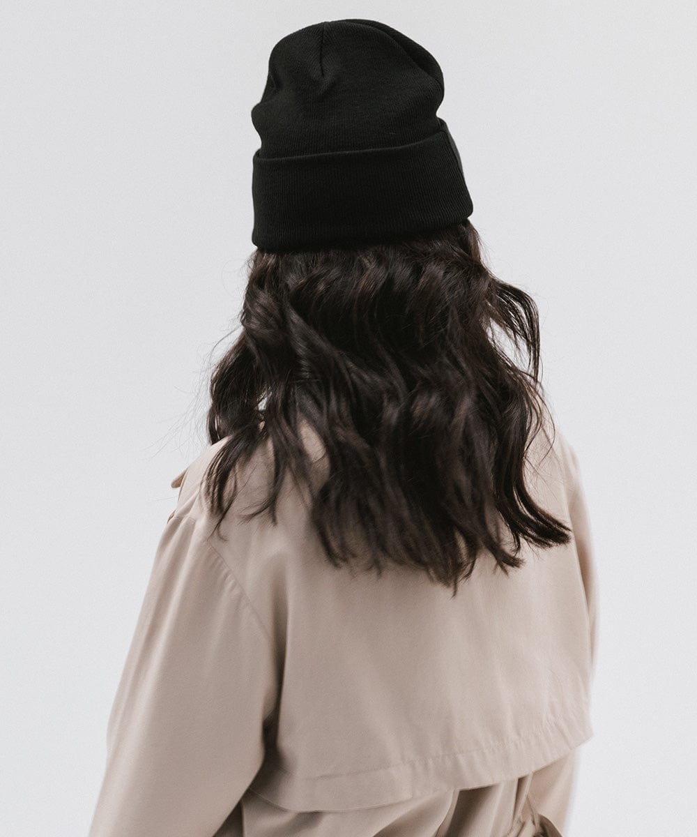 Woman wearing a black knit beanie and a beige jacket, facing behind against a plain background #color_black