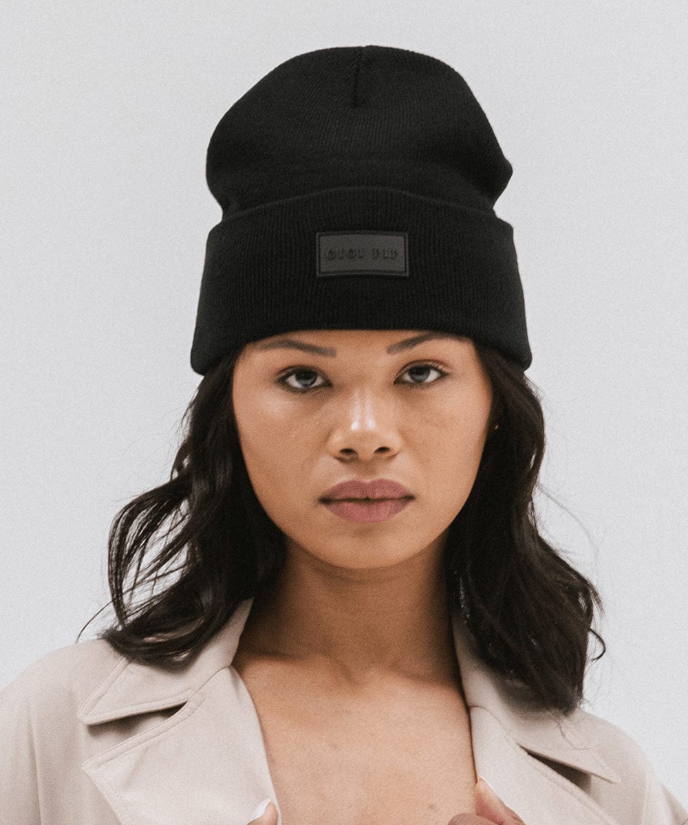 Woman wearing a black knit beanie and a beige coat against a plain background #color_black