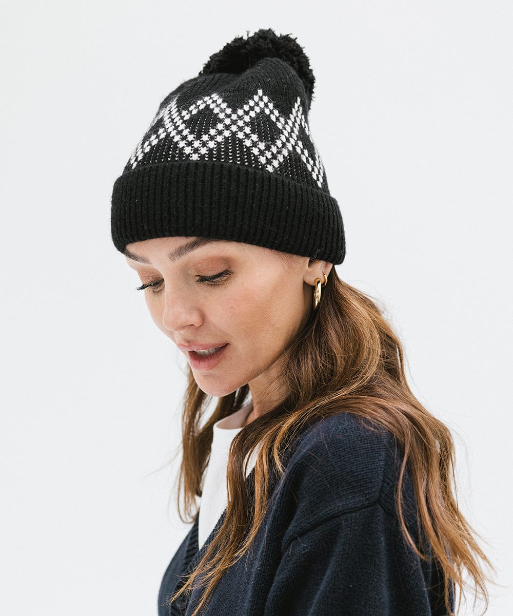 Woman wearing a black knit beanie with a pom-pom and a dark blue sweater, facing sideways against a plain background #color_black