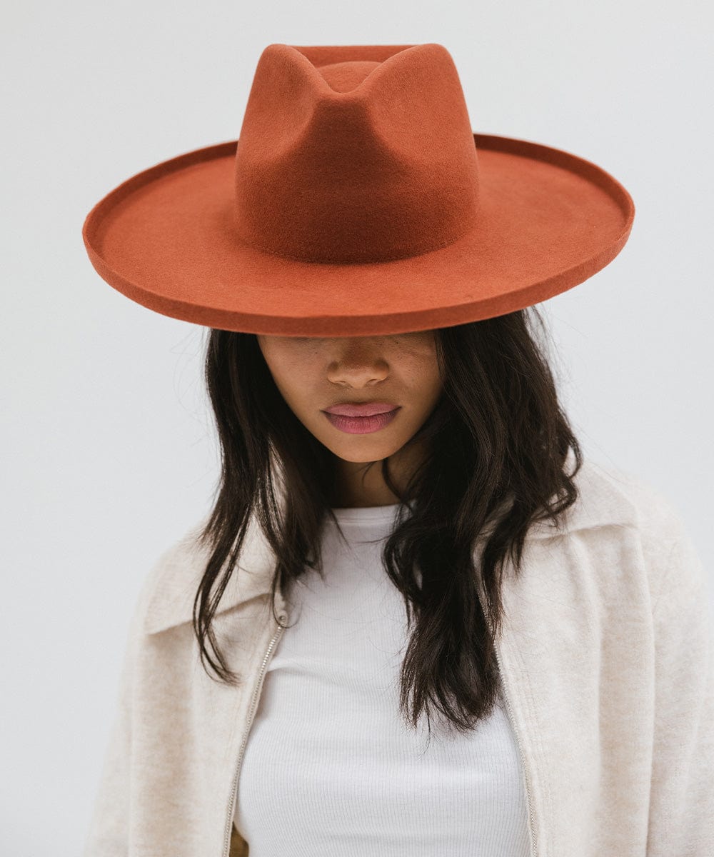 Woman wearing a clay colored pencil brim fedora hat with a wide brim and a white top with a cream jacket against a white background #color_clay