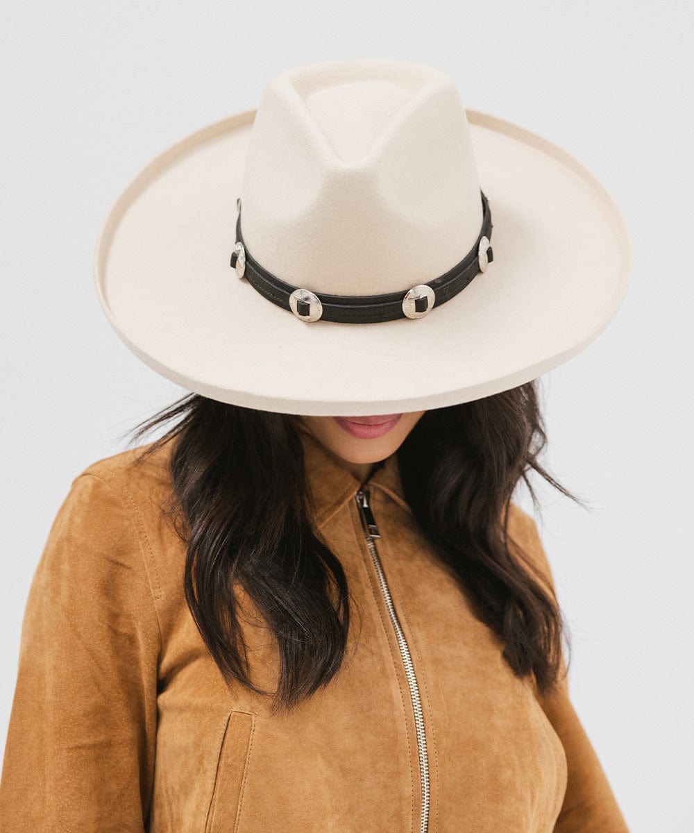 Woman wearing an off white colored pencil brim fedora hat with a wide brim and a hat band, and a brown jacket against a white background #color_off white