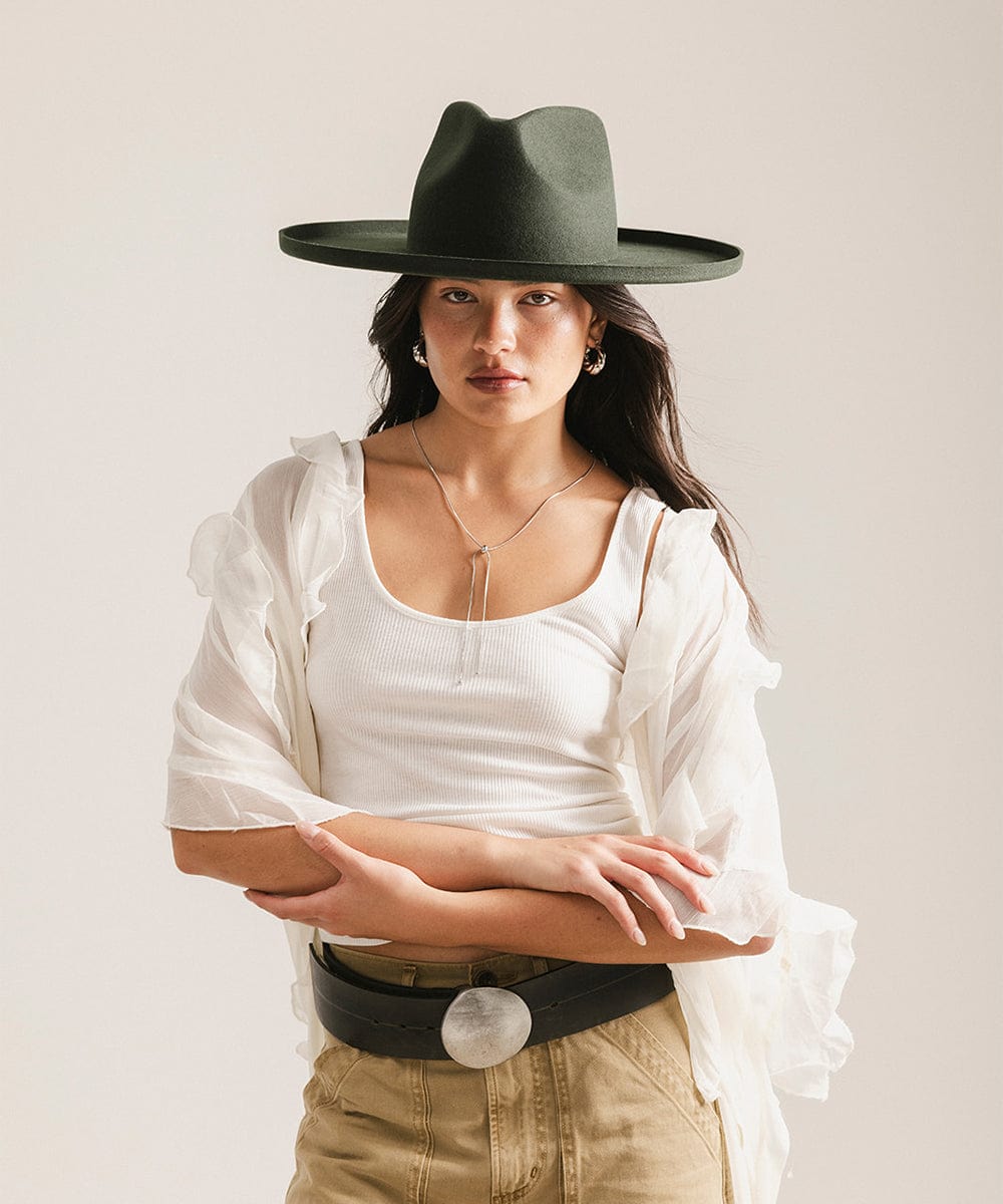 Woman wearing a dark green pencil brim fedora hat and a white top with a white blouse against a plain background #color_dark green