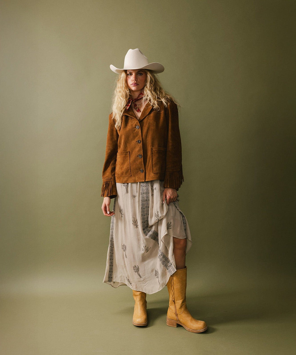 Woman wearing an off white cowboy hat, a cream and grey dress with a brown jacket and yellow boots against a plain background #color_off white