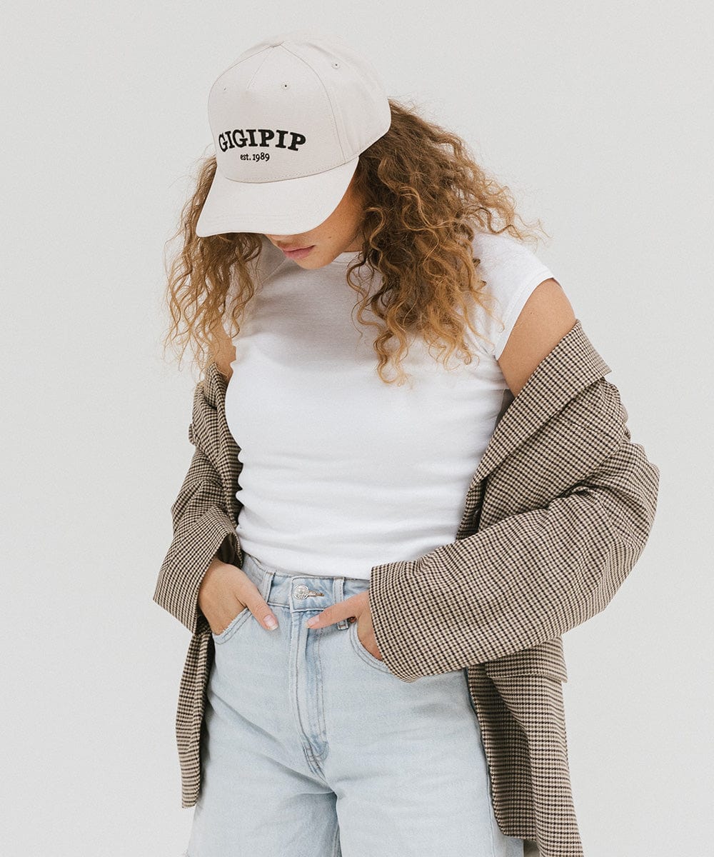 Woman wearing a cream trucker hat with 'GIGI PIP' embroidered in black on the front, a white top with a coat, and blue jeans, posing against a plain background #color_cream