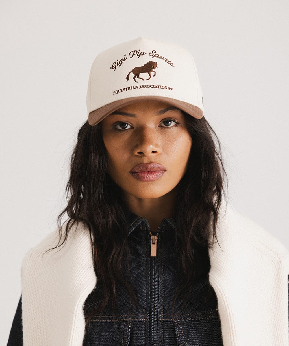 Woman wearing a cream and chocolate brown Trucker hat with 'Gigi Pip Sports' and horse logo and a denim jacket against a plain background #color_cream - chocolate brown