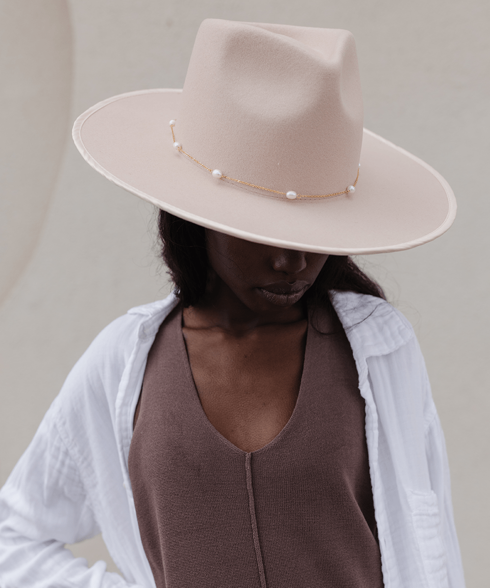 Woman wearing a cream hat with a gold pearl chain band and a brown top with a white shirt against a plain background #color_gold