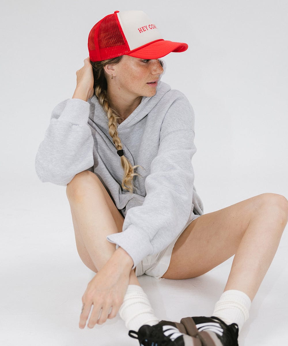 Woman wearing a cream and vintage red cap with 'Hey Cowboy' text and a grey sweater, sitting against a plain background #color_cream - vintage red