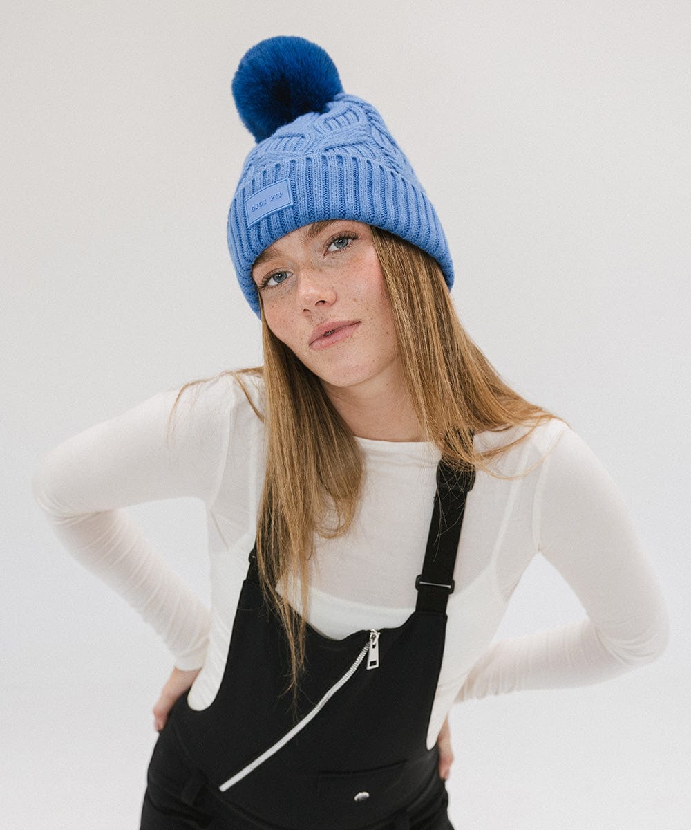 Woman wearing an arctic blue colored knit beanie with a pom-pom and a white t-shirt with a black sleeveless jacket against a plain background #color_arctic blue
