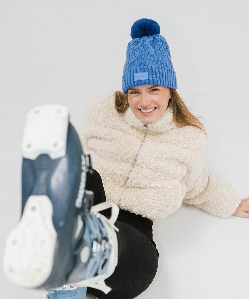 Woman wearing an arctic blue colored knit beanie with a pom-pom, a white sweater, black pants and blue shoes, sitting against a plain background #color_arctic blue