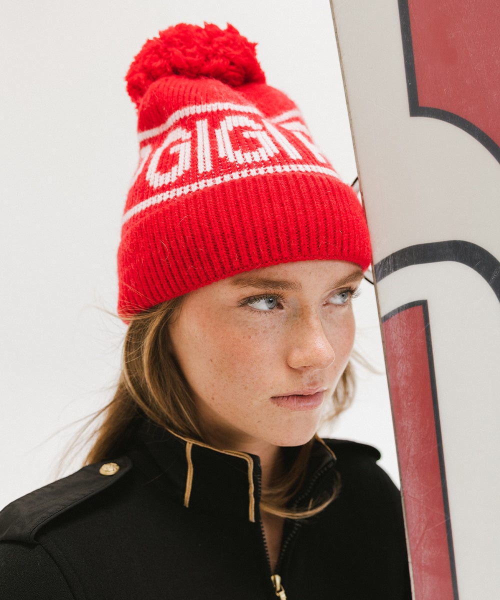 Woman wearing a ruby red colored knit beanie with pom-pom and 'GIGI PIP' branding and a black jacket against a plain background #color_ruby red