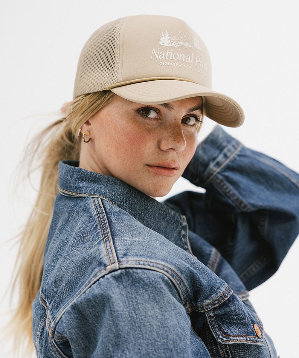 Woman wearing a tan trucker hat with 'National Park' logo and a denim jacket, posing sideways against a plain background #color_tan