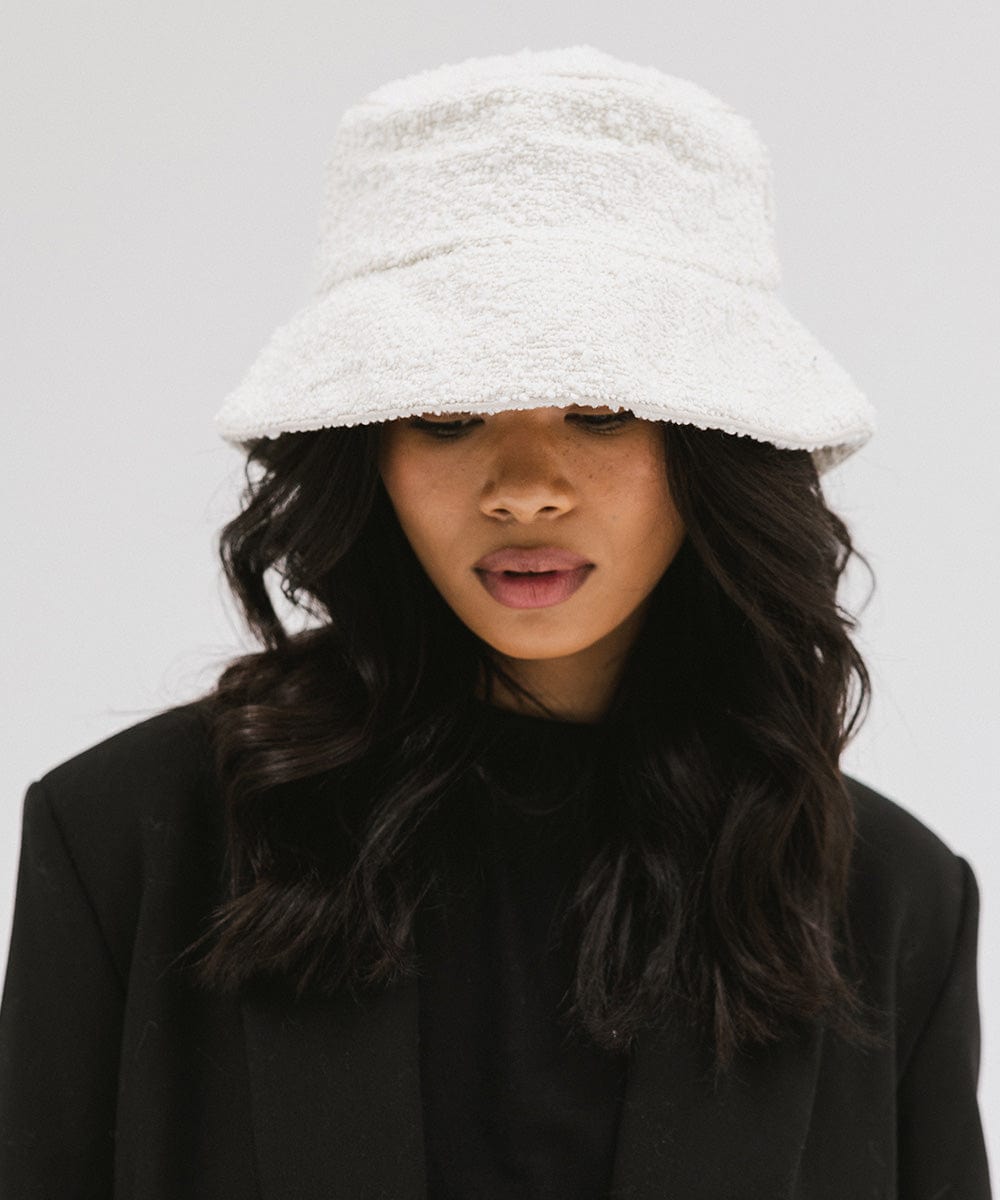 Woman wearing a white bucket hat and a black t-shirt with a black blazer, looking down against a plain background #color_white