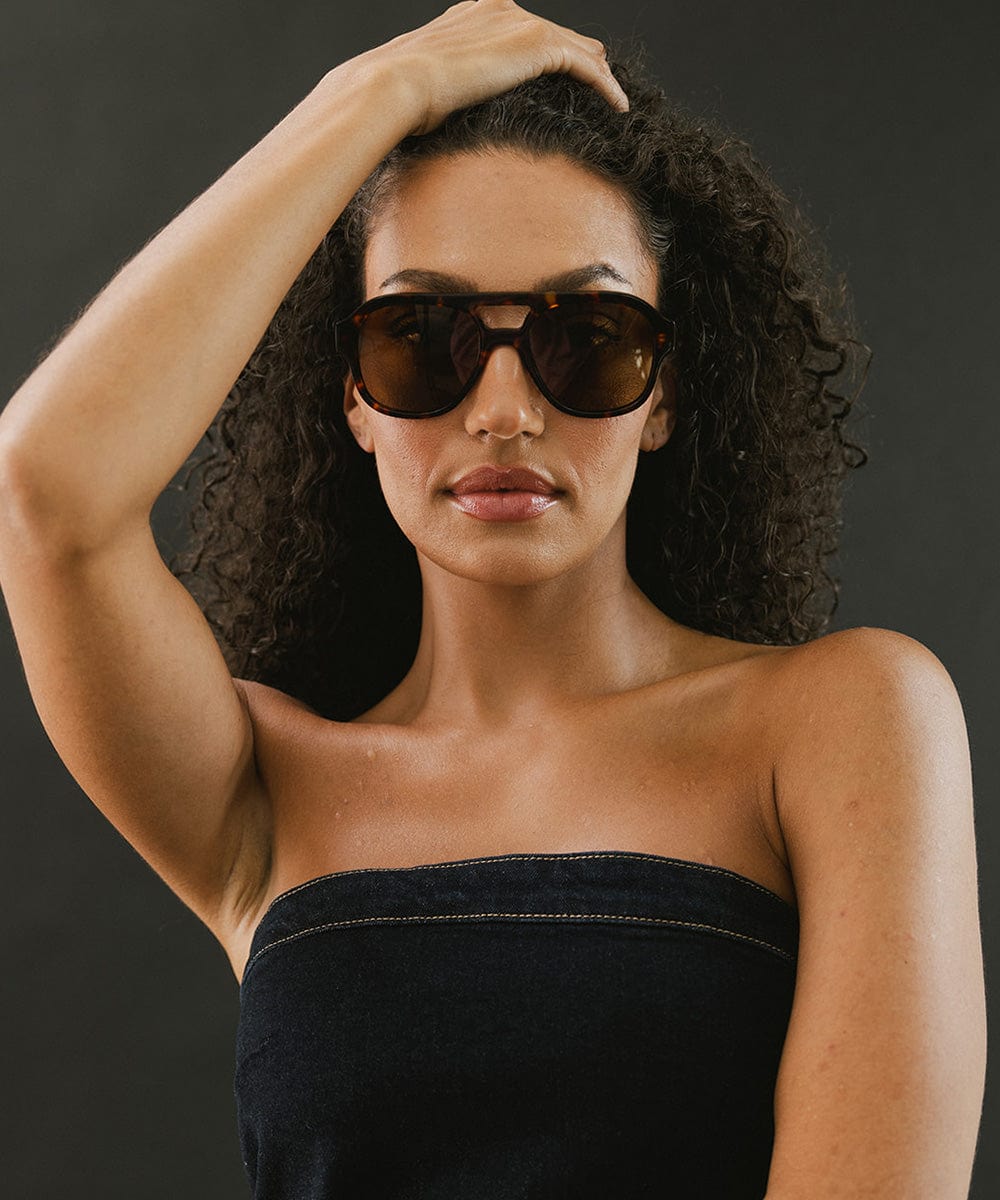 Woman wearing tortoise colored aviator sunglasses and a denim top against a dark grey background #color_tortoise