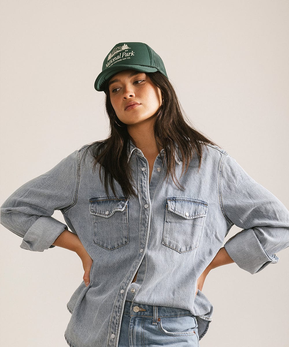 Woman wearing a forest green colored trucker hat with 'National Park' logo and a denim shirt, posing against a plain background #color_forest green