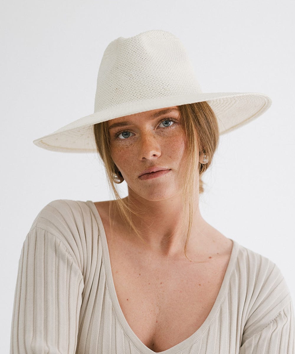 Woman wearing a white wide brim fedora straw hat and a grey top against a plain background #color_white