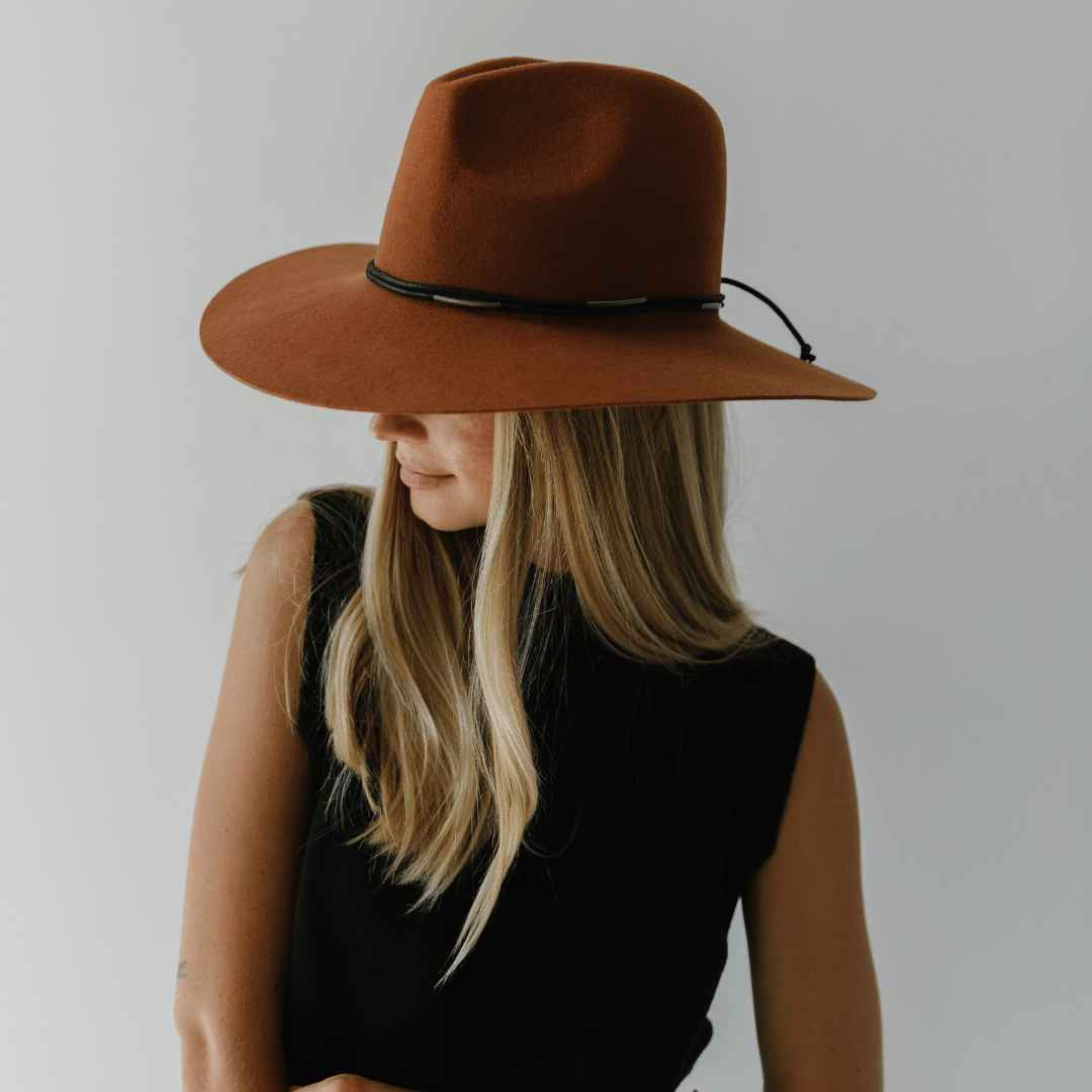 Woman wearing a terracotta colored wide brim hat with a hat band and a black top against a plain background #color_terracotta