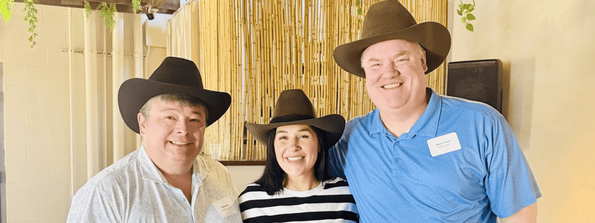 Three people wearing cowboy hats and casual clothing standing together in front of a bamboo screen at a custom hat bar event.