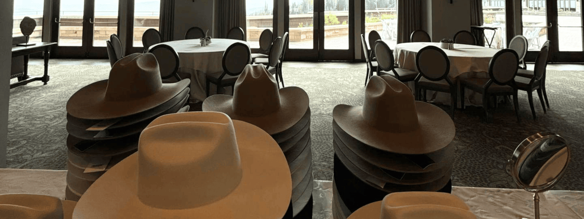 Stacks of wide-brim cowboy hats displayed indoors in a large event space with round tables and tall windows in the background at a custom hat bar event.