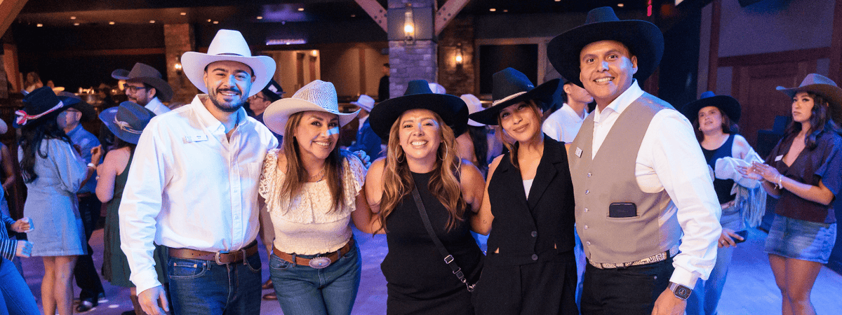A group of five people posing together in a dimly lit indoor event space, dressed in western-style attire including cowboy hats, vests, and jeans at a custom hat bar event.