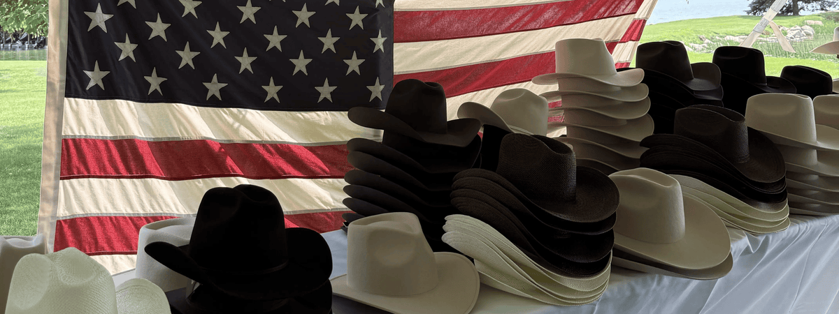 Multiple stacks of cowboy hats in black, brown, cream, and tan are displayed on a table in front of a large American flag, with an outdoor field visible in the background at a custom hat bar event.
