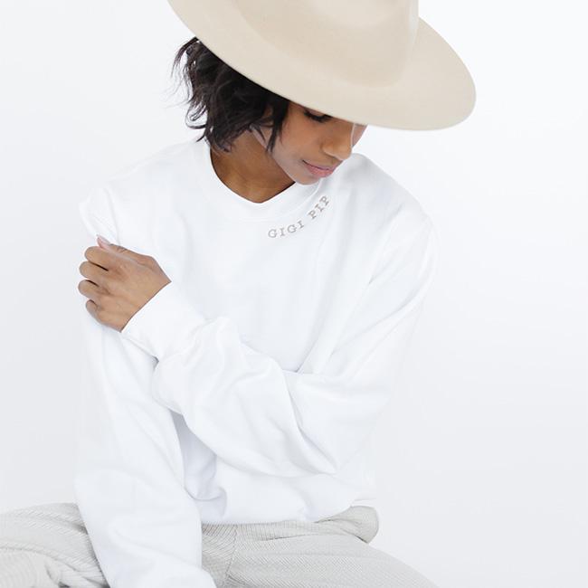 Woman wearing a beige hat, a white sweatshirt with the text 'GIGI PIP' in beige and grey pants, looking down against a plain background #color_white-beige