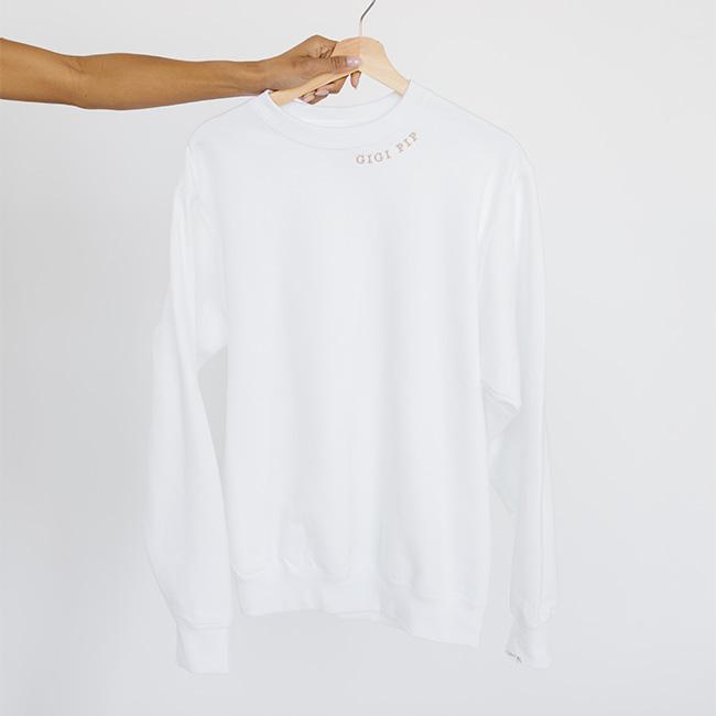 Person carrying a white sweatshirt with the text 'GIGI PIP' in beige  against a plain background #color_white-beige