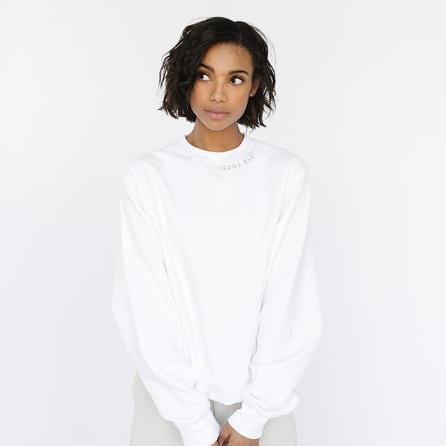 Woman wearing a white sweatshirt with the text 'GIGI PIP' in beige, looking sideways  against a plain background #color_white-beige