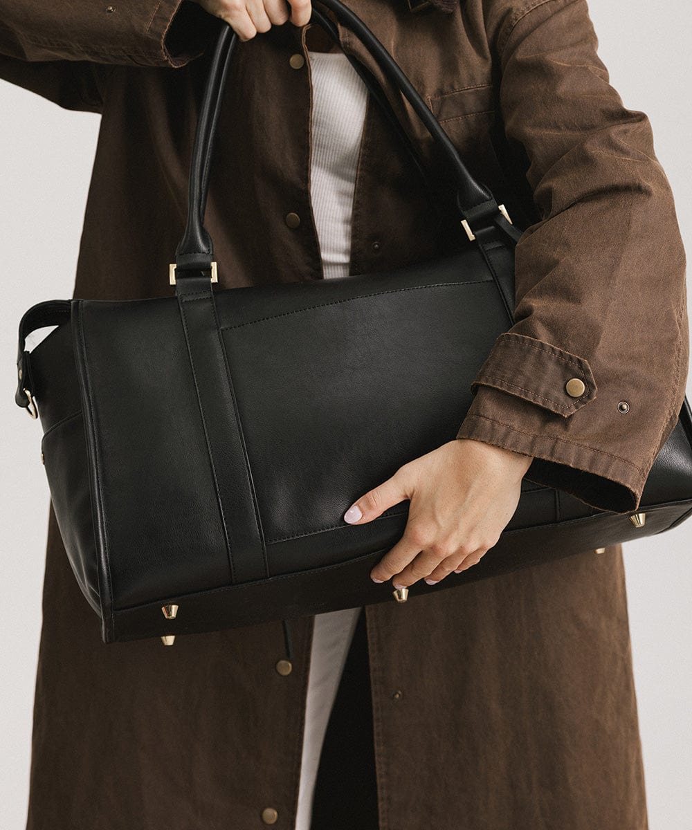 Person wearing a brown coat carrying a black vegan leather weekender bag against a plain background #color_black