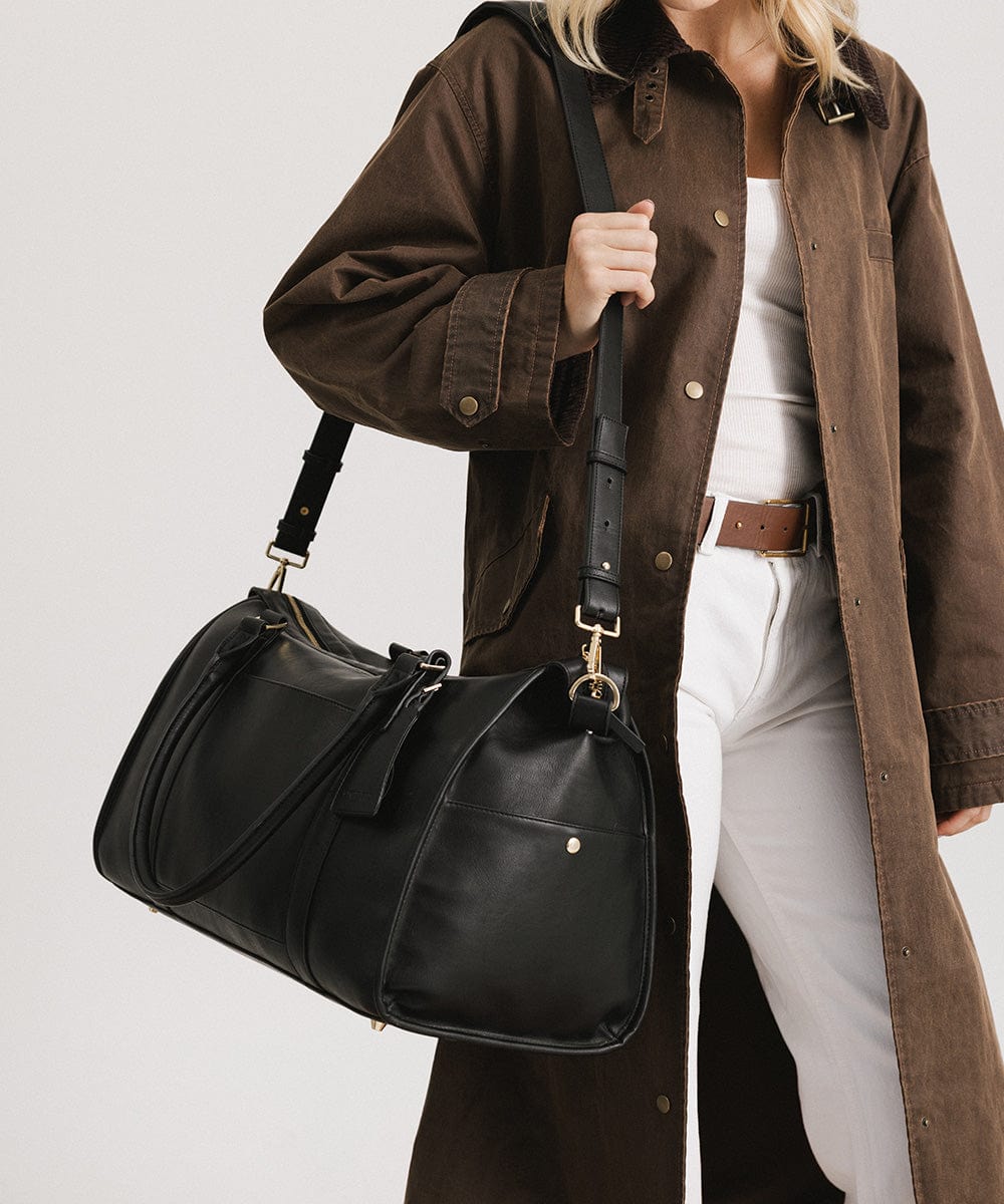 Person wearing a white top with a brown coat and white pants, carrying a black vegan leather weekender bag against a plain background #color_black