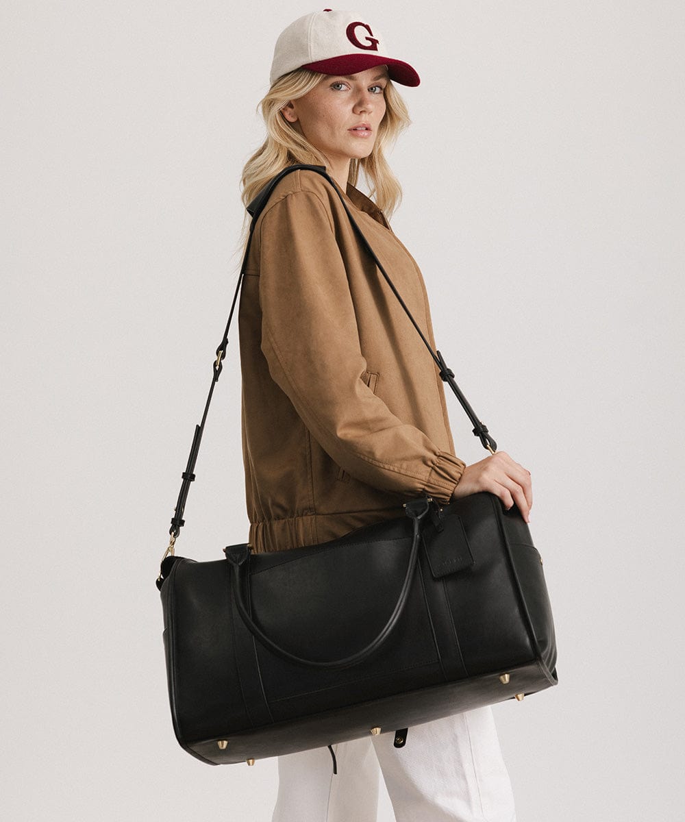 Woman wearing a cap and a brown coat carrying a black vegan leather weekender bag, posing sideways  against a plain background #color_black