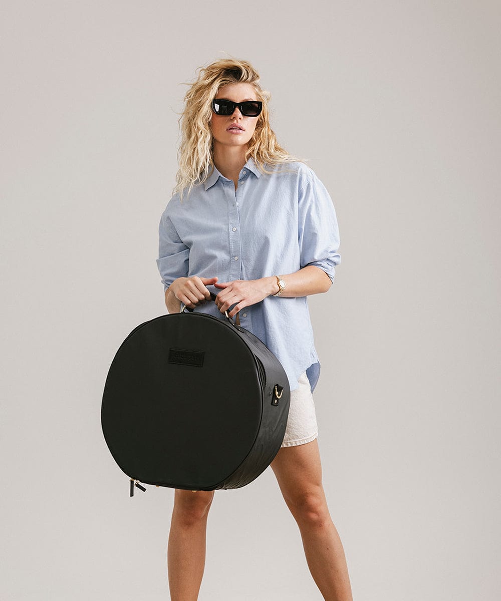 Woman wearing sunglasses, a light blue shirt and white shorts carrying a black round hat travel case against a plain background #color_black
