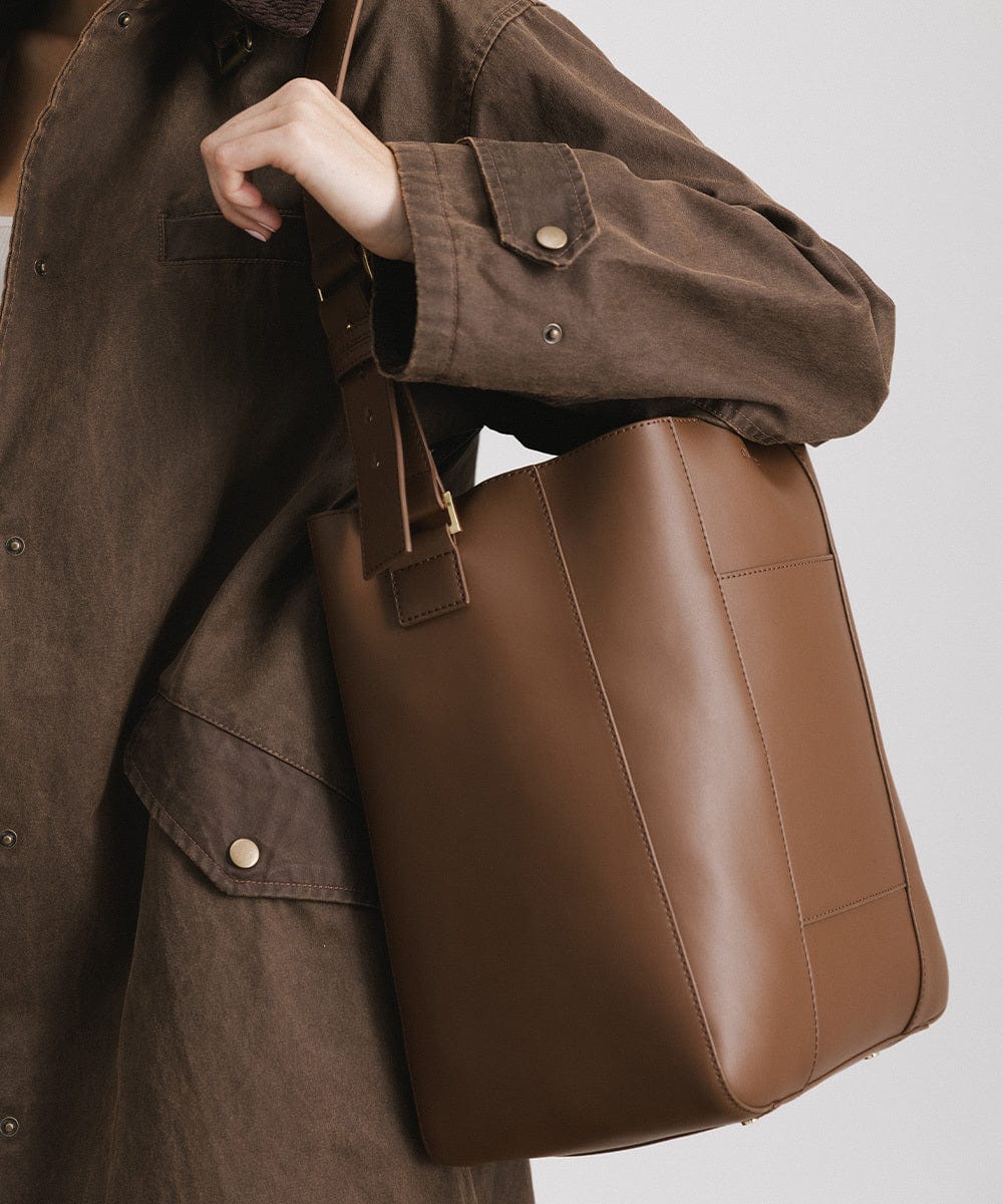 A person wearing a brown coat carrying a chocolate colored Dia Everyday Leather Tote Bag against a plain background  #color_chocolate