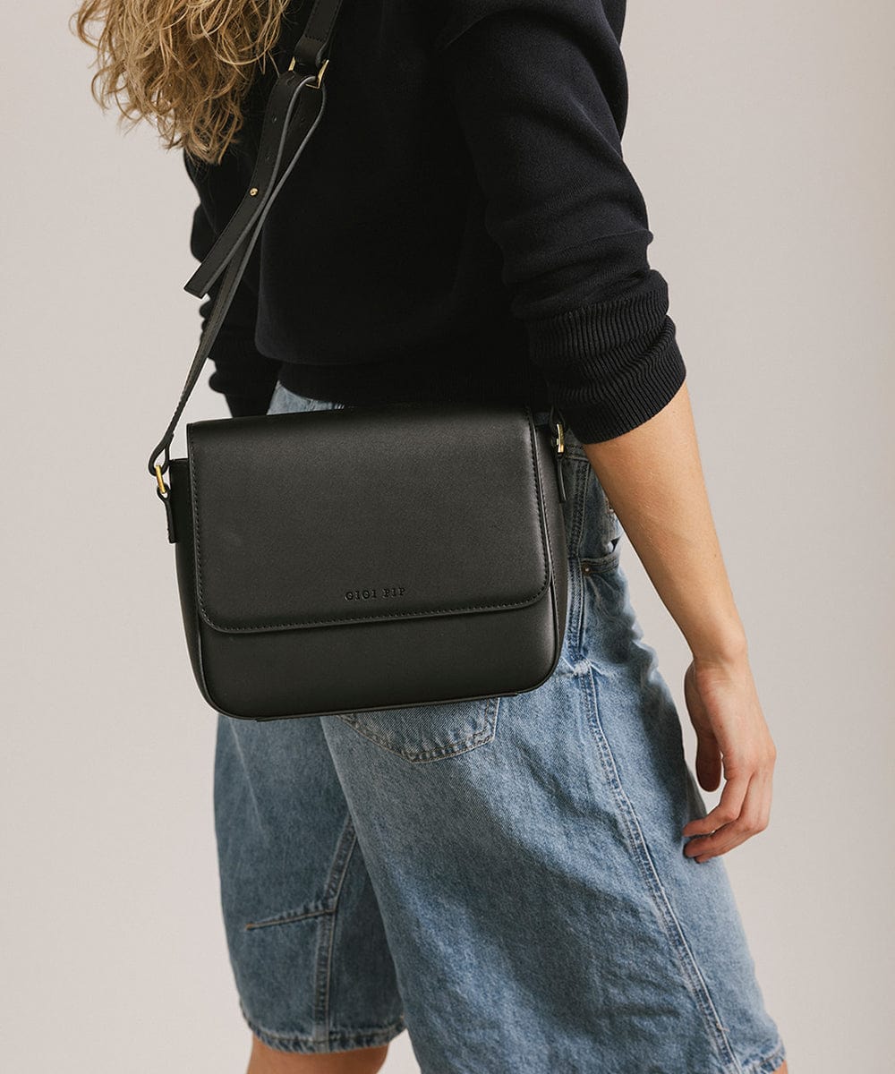 Person wearing a black sweater and denim shorts carrying a black Rhys Crossbody Genuine Leather Bag against a plain background  #color_black