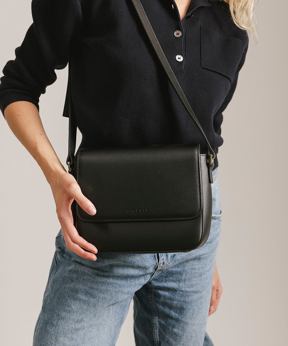 Person wearing a black shirt and blue jeans carrying a black Rhys Crossbody Genuine Leather Bag against a plain background  #color_black