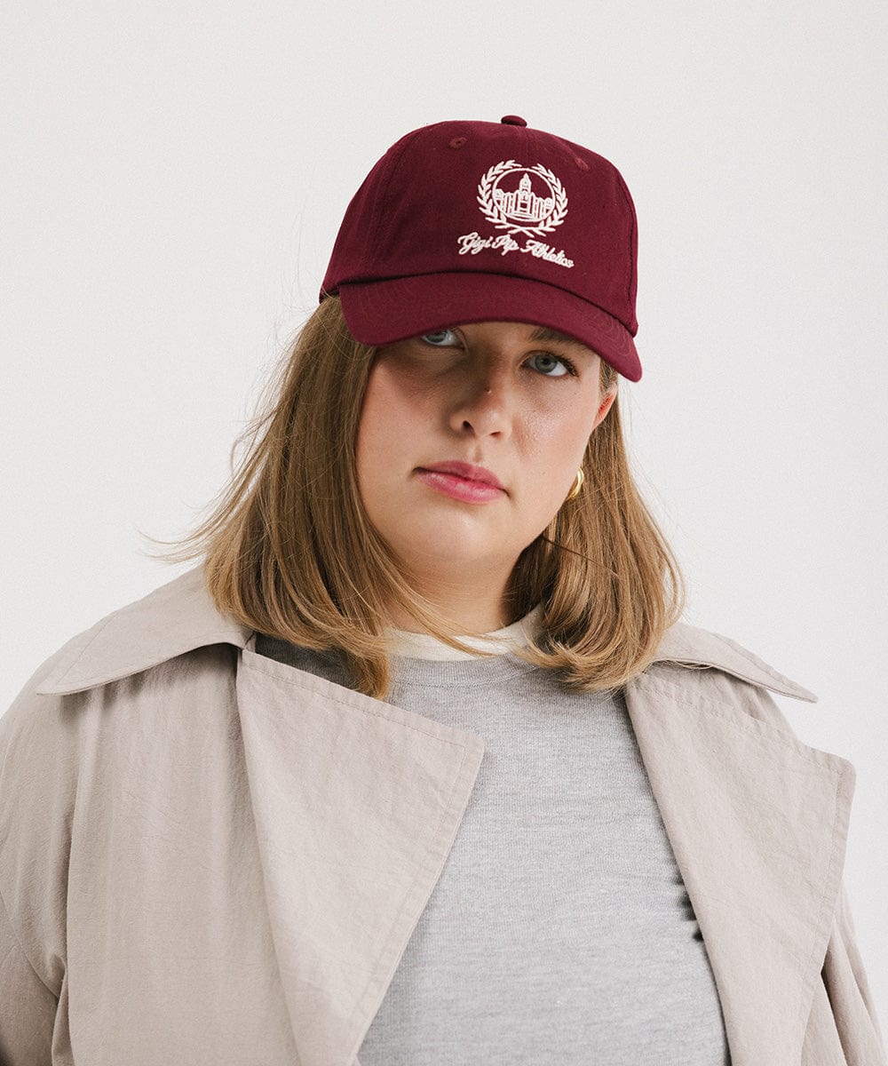Woman wearing a burgundy ball cap with a white logo and text and a grey t-shirt with a beige shirt against a plain background  #color_burgundy