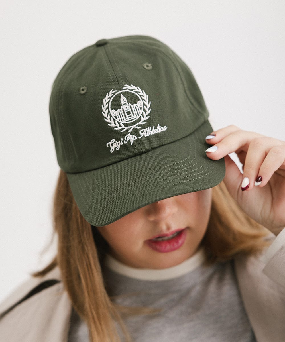 Woman wearing a dark green ball cap with a white logo and text and a grey shirt against a plain background  #color_dark green