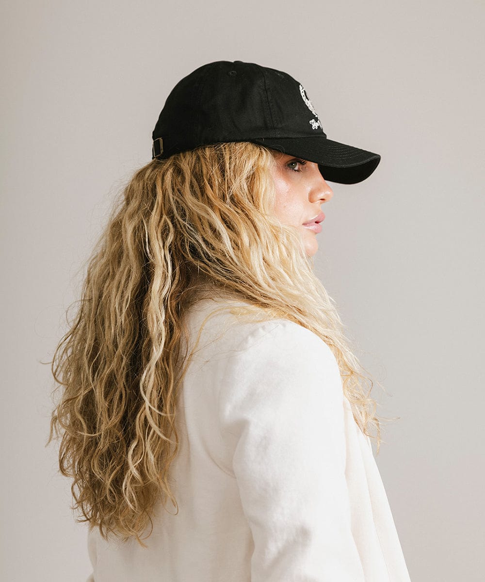 Woman wearing a black ball cap with a white logo and text and a white blazer, facing sideways against a plain background  #color_black