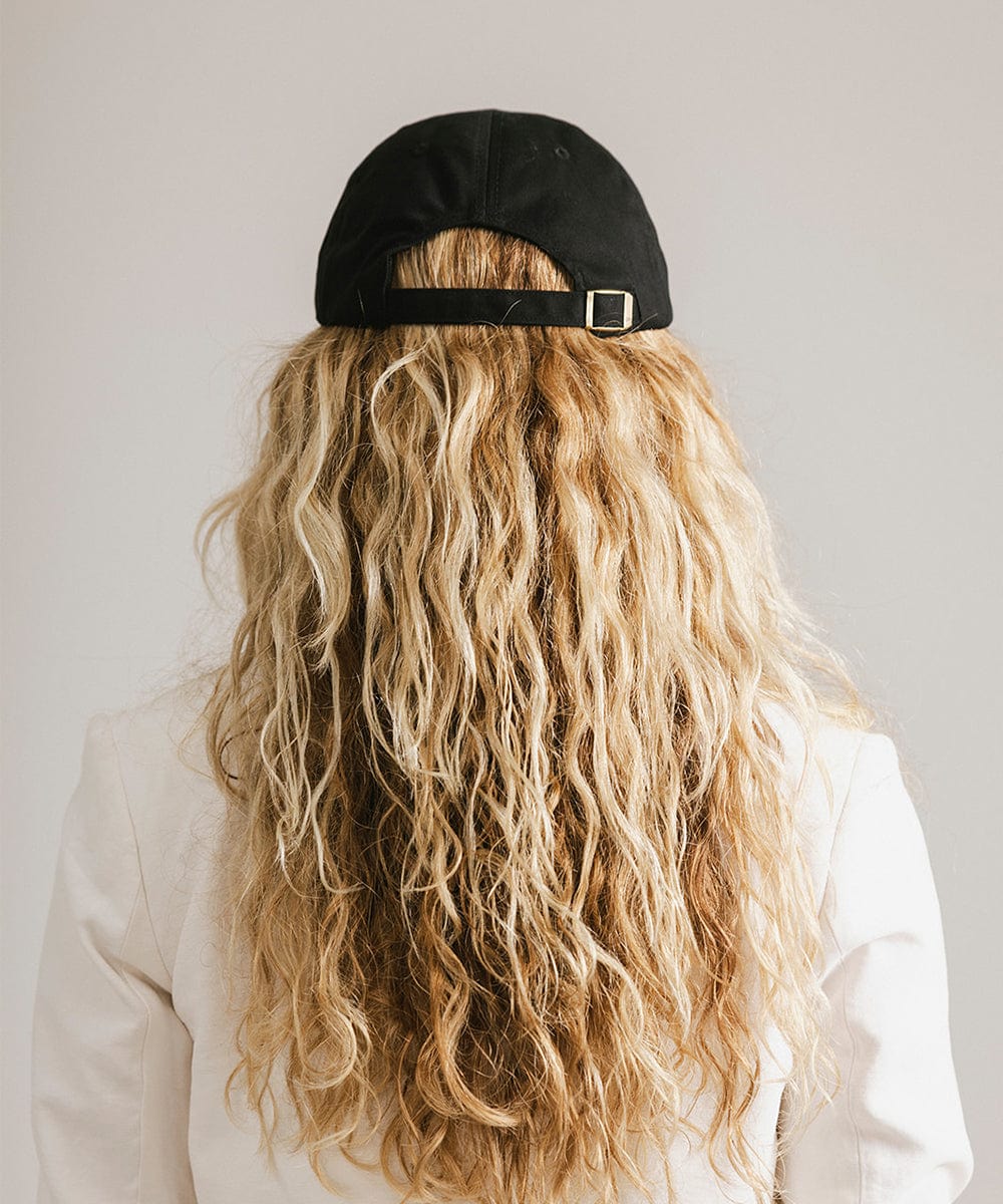 Woman wearing a black ball cap and a white blazer, facing behind against a plain background  #color_black