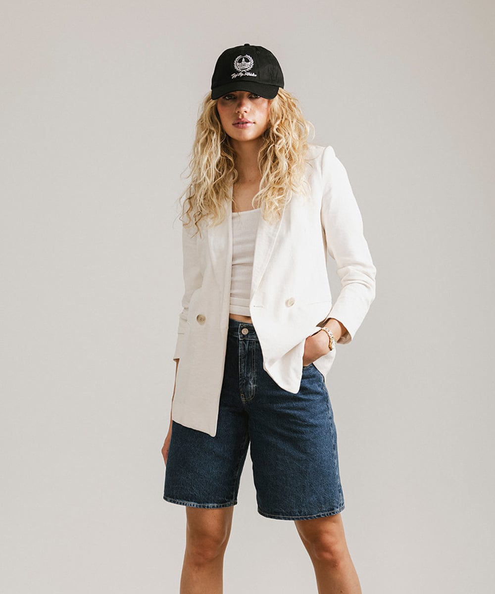 Woman wearing a black ball cap with a white logo and text, a white top with a white blazer and denim shorts against a plain background  #color_black