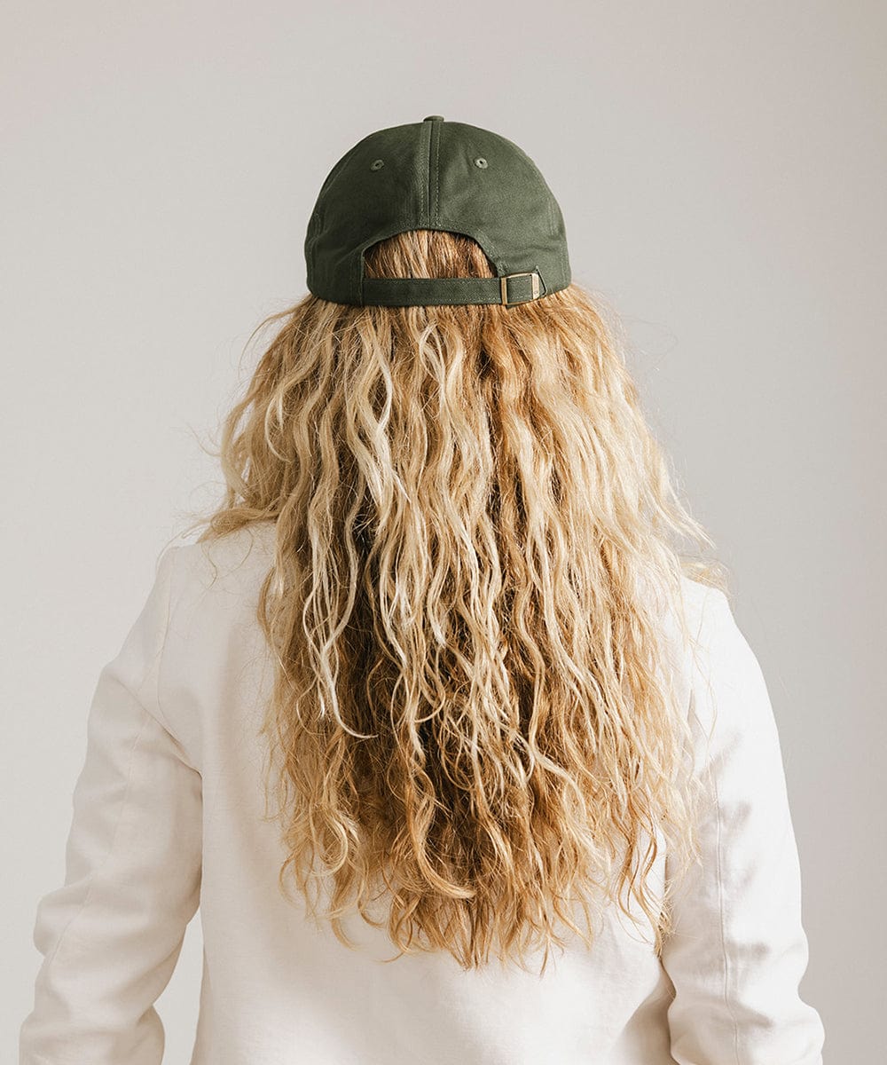 Woman wearing a dark green ball cap   and a white blazer, facing behind against a plain background  #color_dark green