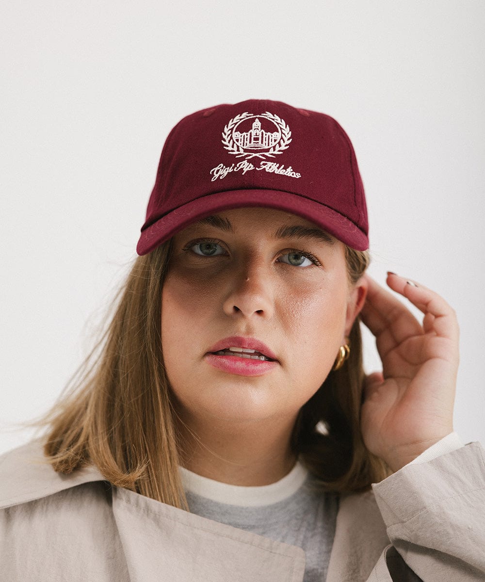 Woman wearing a burgundy ball cap with a white logo and text against a plain background  #color_burgundy